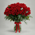 A large bouquet of red roses with green foliage and a red ribbon tied around a clear glass vase, photographed against a light gray studio background.