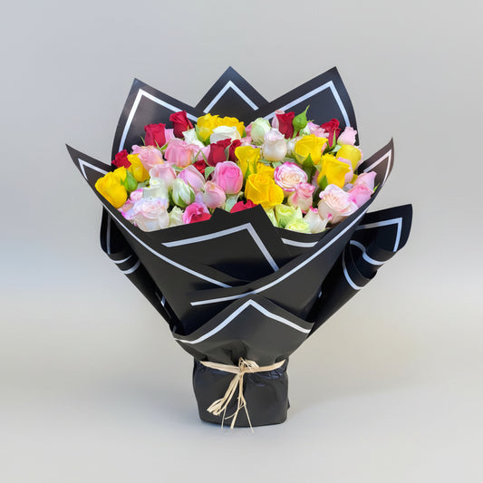 A bouquet of yellow, pink, red, and white flowers wrapped in black paper with decorative white lines and a rustic tie at the base.