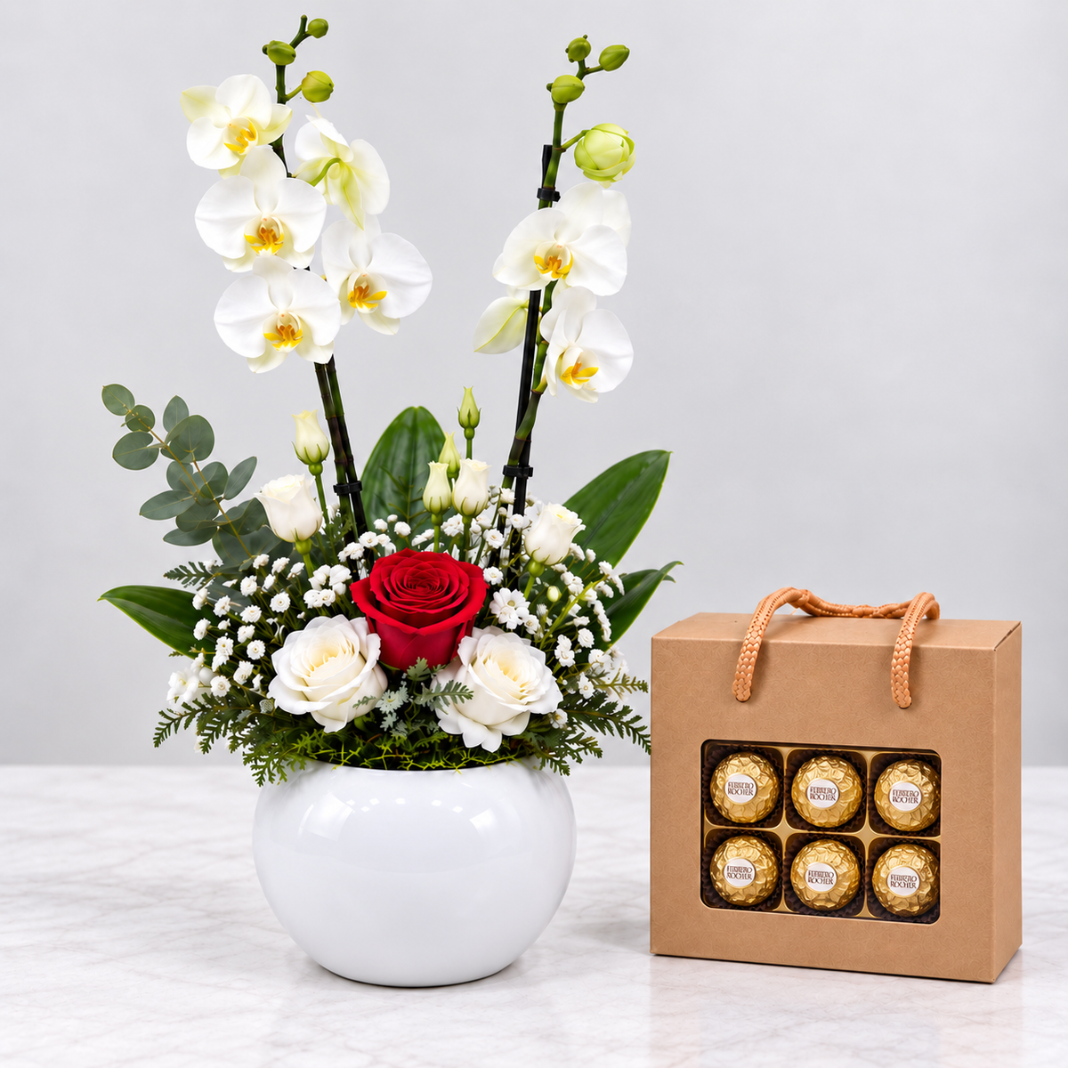 A floral arrangement in a white round vase with white orchids, white roses, green foliage, and a single red rose, placed beside a kraft gift box containing six Ferrero Rocher chocolates.