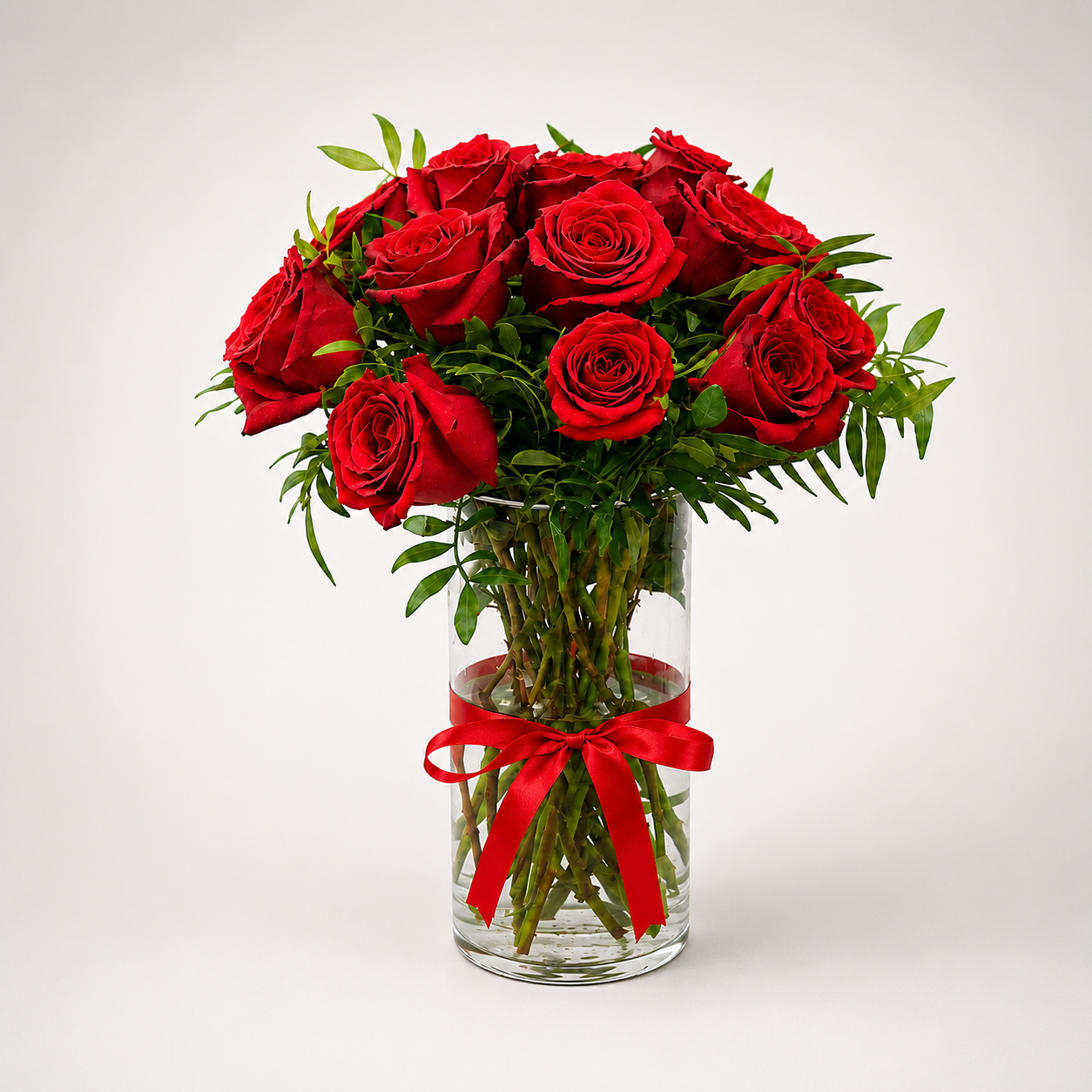10 premium red roses arranged in a clear glass cylinder vase with green filler foliage and a red satin ribbon bow"