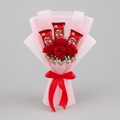 A novelty bouquet made from three KitKat chocolate bars arranged like flowers, with a single red rose in the center, small white baby's breath accents, pink tissue paper wrapping, and a red ribbon bow, photographed on a light gray background.